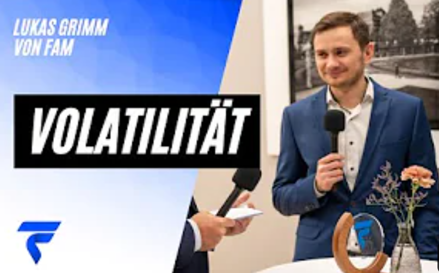 Ein Mann im Anzug spricht in ein Mikrofon, daneben steht &quot;Volatilität&quot;. Links daneben Schrifteinblendung: &quot;Lukas Grimm von FAM&quot;. Auf einem Tisch stehen Blumen und ein dekoratives Objekt.