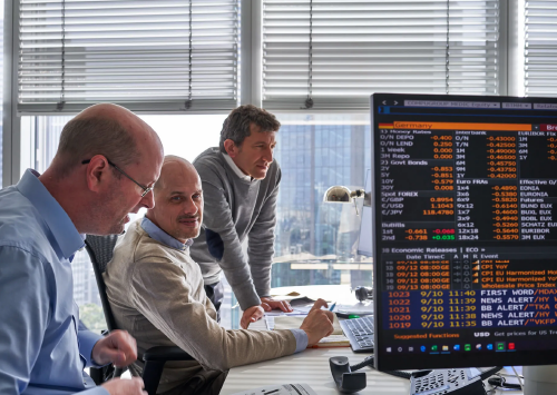 Drei Männer analysieren Finanzdaten auf einem Computerbildschirm in einem modernen Büro. Sie diskutieren über Aktienkurse und Wirtschaftsinformationen. Im Hintergrund sind große Fenster mit Stadtblick.