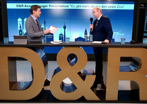 Zwei Männer führen eine Diskussion auf einer Bühne mit einem großen D&amp;R-Logo im Vordergrund. Der Bildschirm im Hintergrund zeigt den Titel „Es gibt mehr als nur den einen Zins“ und eine Stadt-Silhouette.