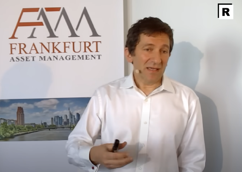 Mann hält Vortrag vor einem Poster von &quot;Frankfurt Asset Management&quot;. Er trägt ein weißes Hemd und benutzt ein Mikrofon. Im Hintergrund ist ein Bild der Frankfurter Skyline. Veranstaltung wirkt professionell und geschäftlich.