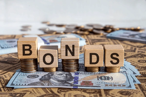Holzklötze mit dem Wort „BONDS“ stehen auf Stapeln von Münzen und US-Dollarnoten. Im Hintergrund liegen weitere Geldscheine und Münzen, symbolisch für Investitionen in Anleihen und Finanzmärkte.