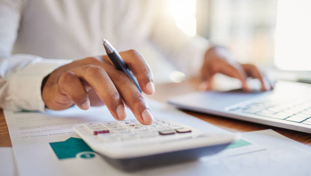 Eine Person in Businesskleidung arbeitet an einem Schreibtisch mit einem Taschenrechner und Laptop. Sie überprüft finanzielle Dokumente. Büro- und Finanzmanagement-Konzept. Fokus auf Hand und Stift, zukunftsorientiertes Arbeiten im Unternehmen.